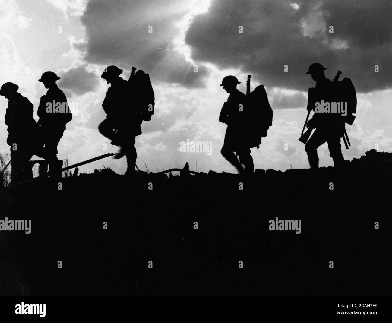 Five soldiers silhouetted against the sky. Rays of sun burst through dark clouds to create a dramatic and atmospheric shot. They are all wearing steel helmets, and three of them are clearly carrying rifles and backpacks. World War I Stock Photo