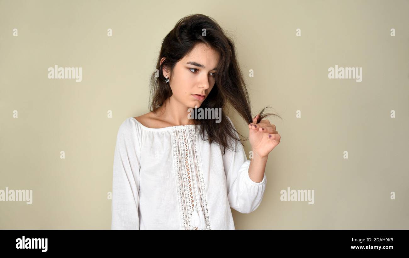 Sad woman with long dry unruly hair looking at split ends of her hair on gray background. Damaged hair concept Stock Photo