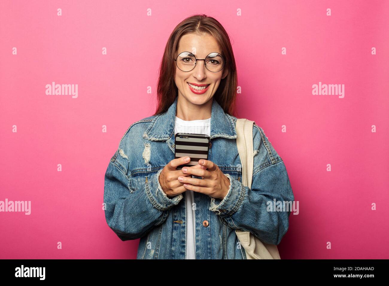 Smiling young woman wearing glasses and denim jacket with phone on pink background. Stock Photo