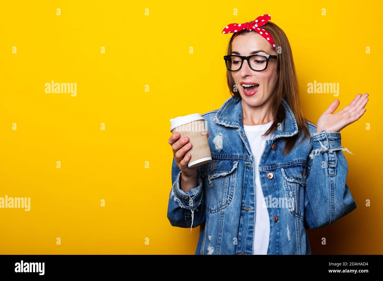 Young woman in denim jacket, headband and glasses looks with surprise at paper cup on yellow background Stock Photo