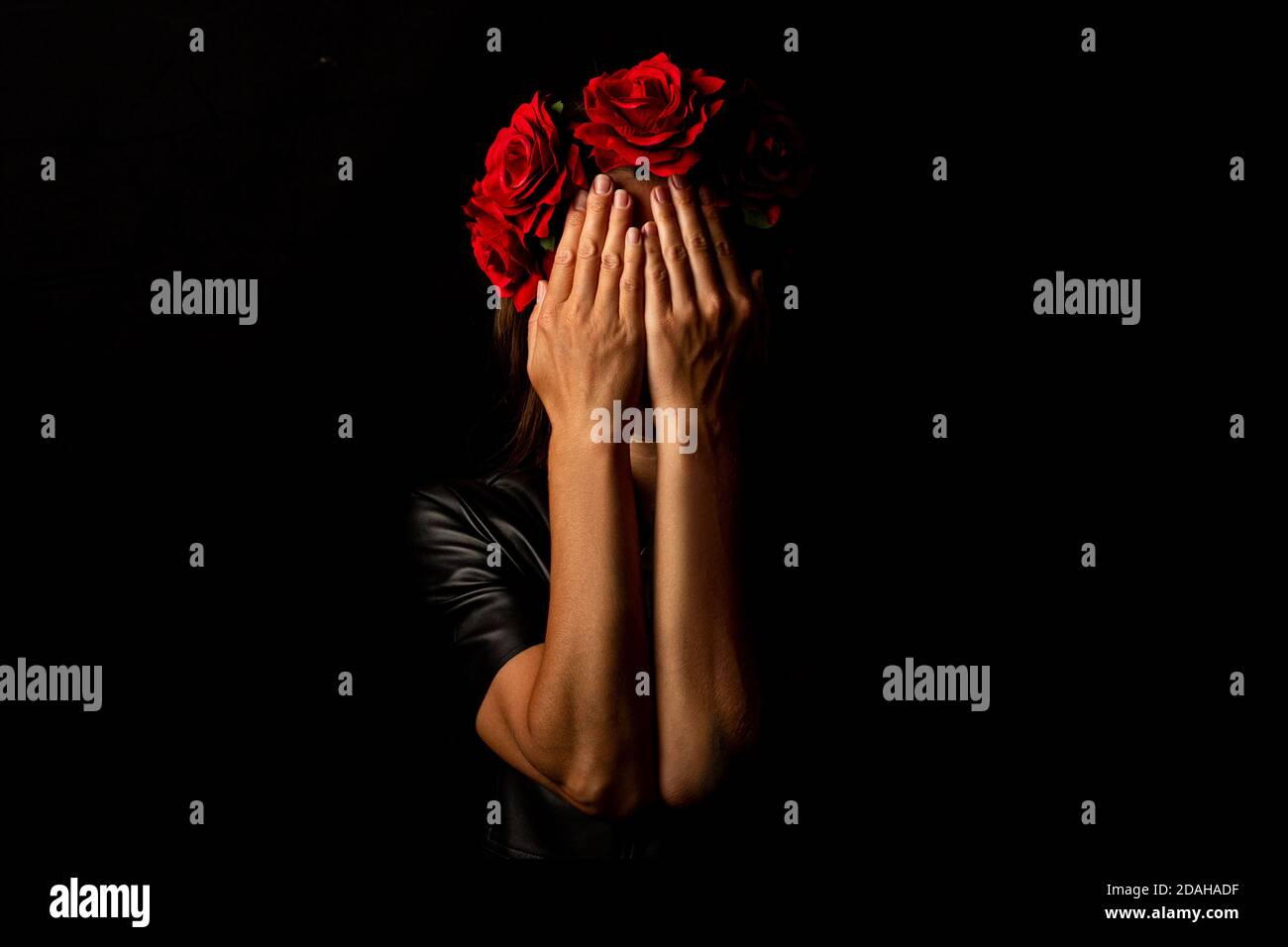 Young woman in a wreath of flowers, covering her face with her palms on a black background. Stock Photo