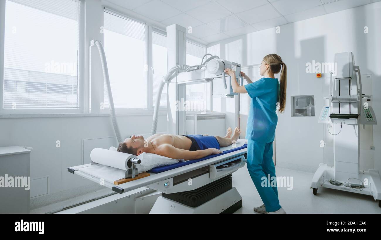 In the Hospital, Low Angle Shot of Man Lying on a Bed, Female Technician adjusts X-Ray Machine. Scanning for Fractures, Broken Limbs, Injuries, Cancer Stock Photo