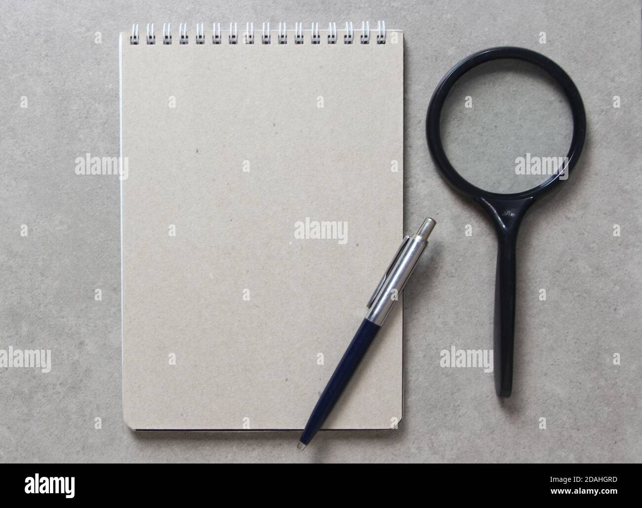 A spring notebook with a sheet of craft paper A5 with a ballpoint pen and black magnifying glass on light grey concrete background. Concept of new idea, business plan and strategy, development and implementation of content. Empty space for text. Stock Photo