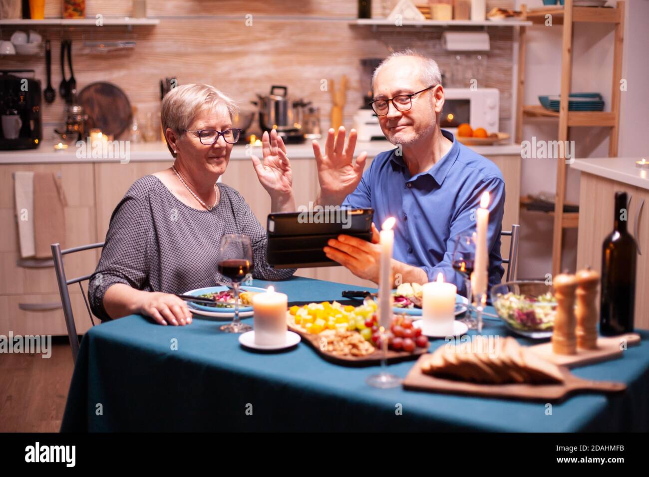 Mature man and woman waving at tablet pc during video conference in kitchen celebrating relationship. Couple sitting at the table, browsing, talking, using internet, celebrating their anniversary in dining room. Stock Photo