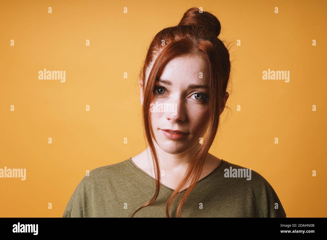 cool young woman with red hair messy bun hairstyle and loose strands at the front against yellow orange color background with opy space Stock Photo