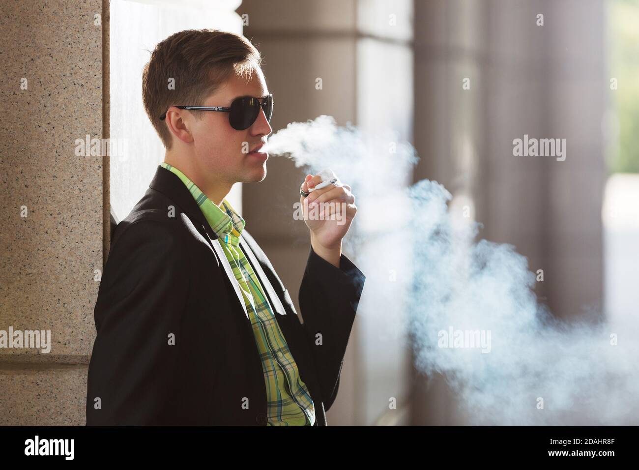 Young man in black blazer and sunglasses smoking a cigarette on city street Stock Photo