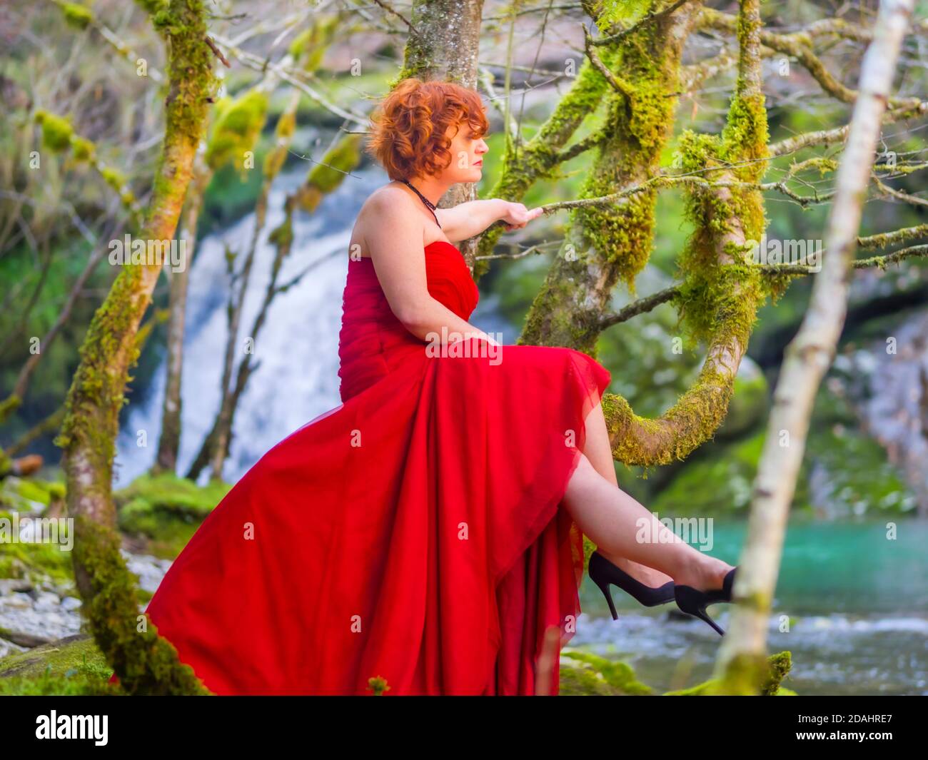 Redhaired curly hair woman in nature before watefall sitting on tree wearing long bright Red dress Black high-heels shoes waiting wait serious Stock Photo