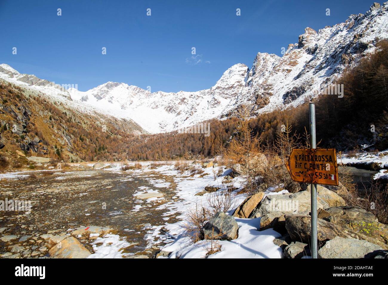 Informative sign and Mountain view from the Preda Rossa Valley in late autumn, with snow and orange pines on a sunny day. Val Masino, Lombardy, Italy. Stock Photo