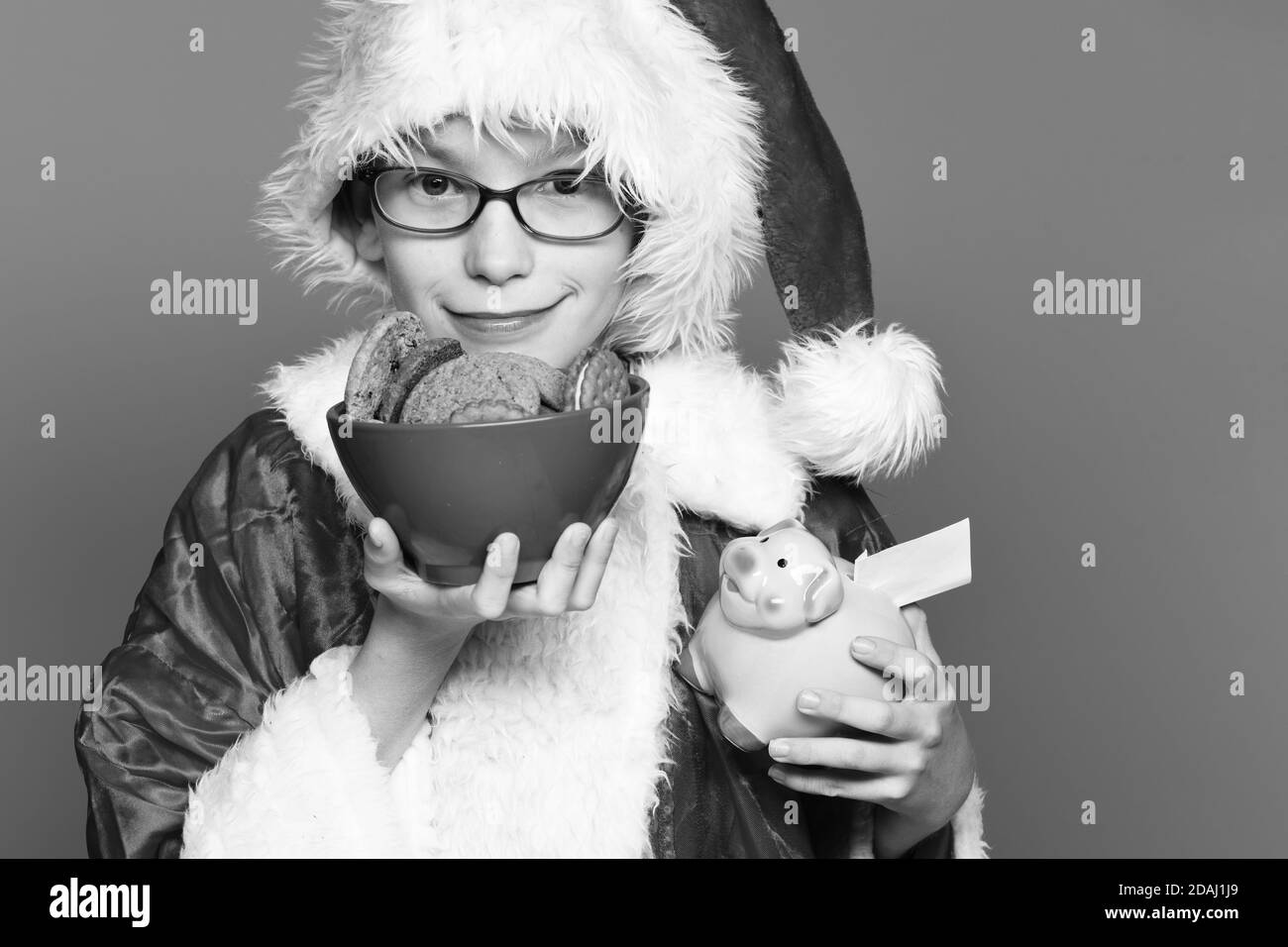 young cute santa claus boy with glasses in red sweater and new year christmas hat holding pink piggy pig bank and chocolate hip cookies in bowl, on blue studio background Stock Photo