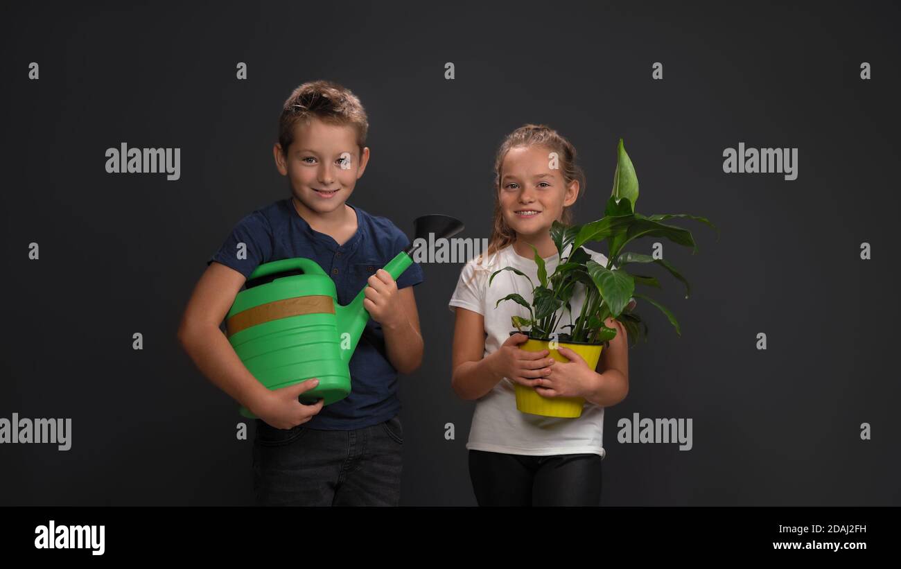 Laughing Caucasian teenagers, a boy holding a watering can, a girl holding a plant in a flowerpot. Isolated on dark gray background. Plant care Stock Photo