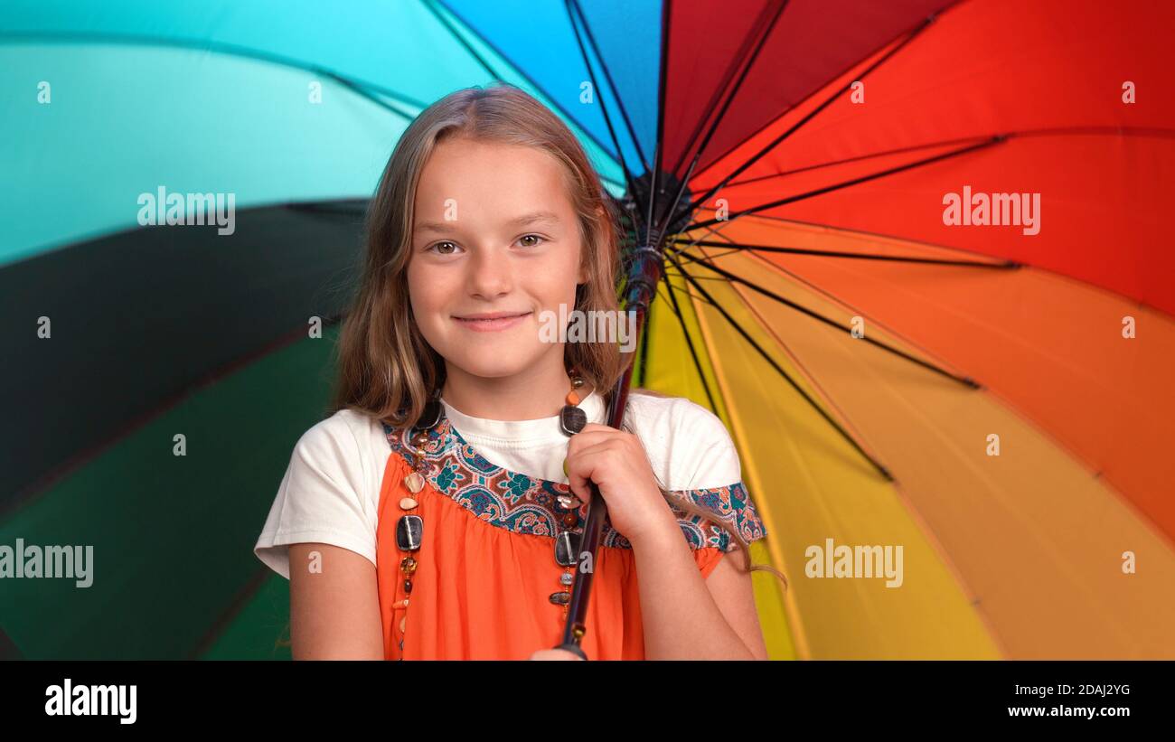 Smiling girl in mother's sundress with beads holds open bright multi-colored umbrella. Caucasian child looks at camera and smiles Stock Photo