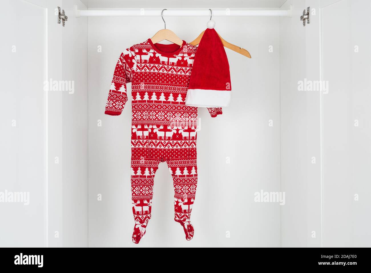 Horizontal color image with a front view of a white and red christmas hat and pajamas hanged on a wooden hangers in the middle of a white closet. Stock Photo