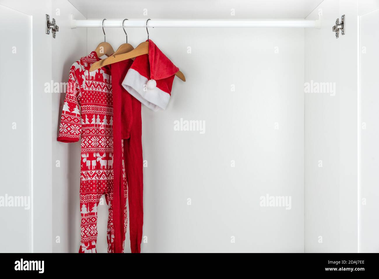 Horizontal color image with a front view of a white and red christmas pajamas, hat and tights hanging on a wooden hangers on the left of a white close Stock Photo