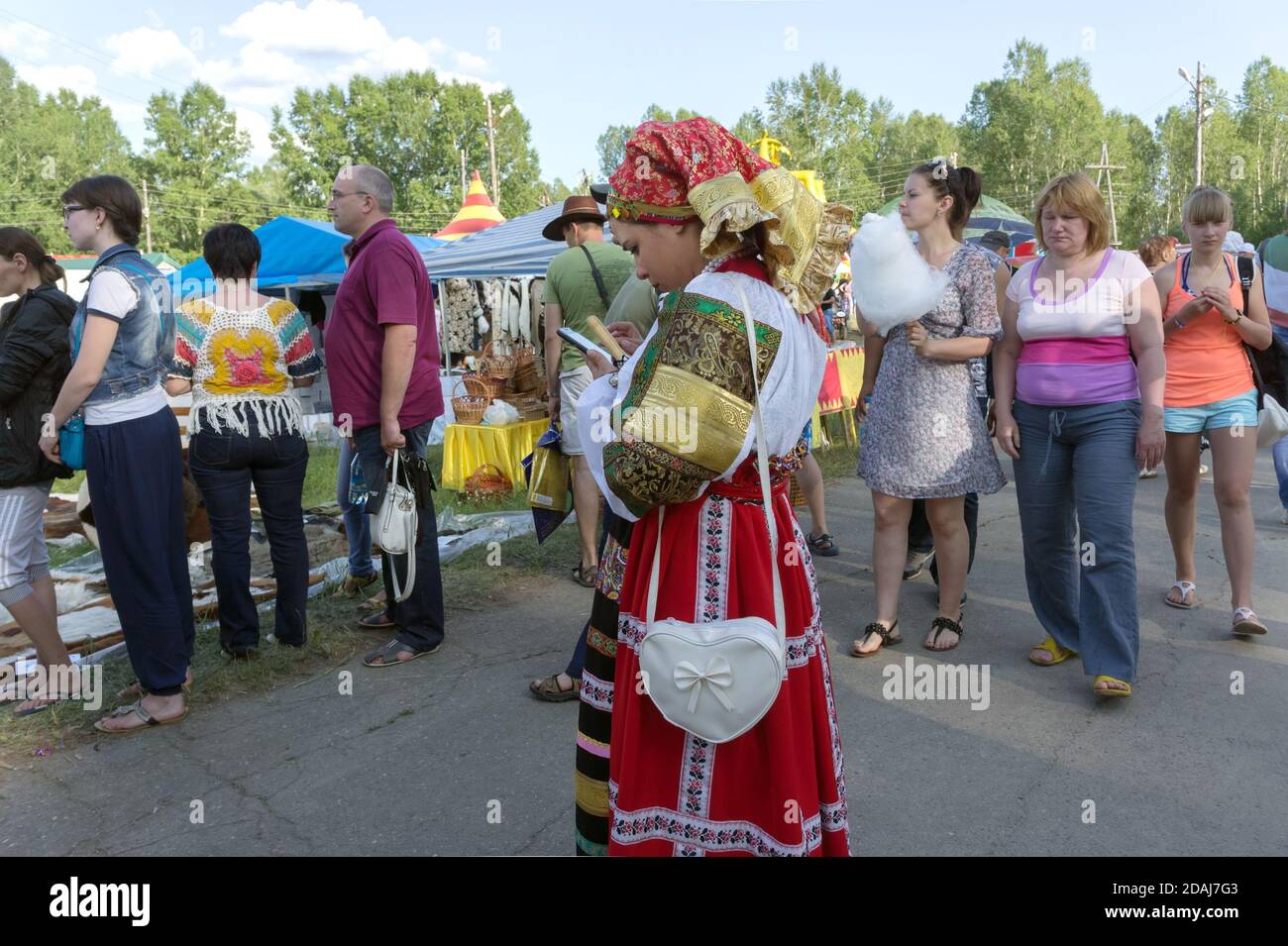 Girl in folk costume is standing and looking at a cell phone among the crowd  during the annual Intl festival of music and crafts World of Siberia. Stock Photo