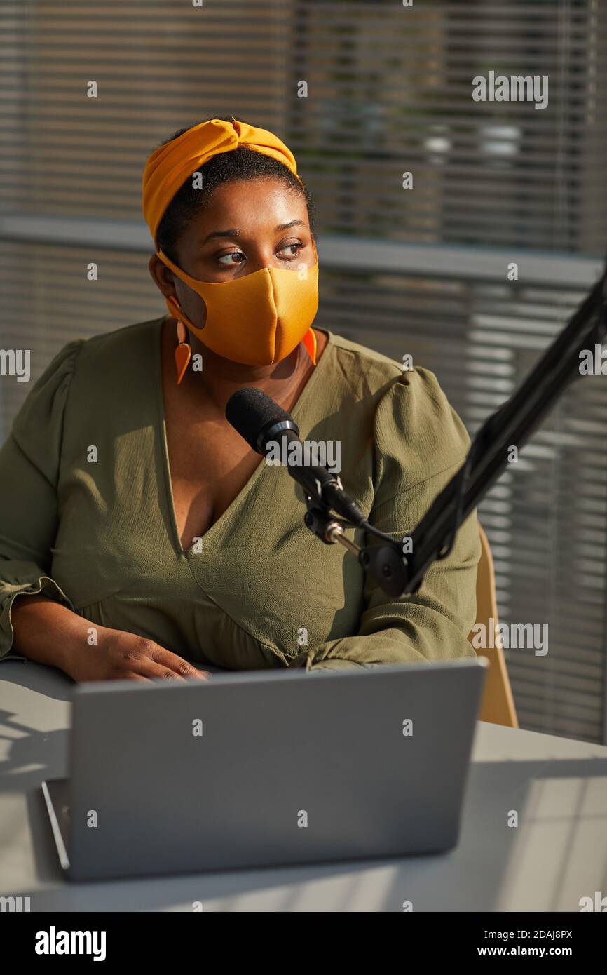 African female interviewer in protective mask using laptop and speaking on microphone during broadcast on radio Stock Photo