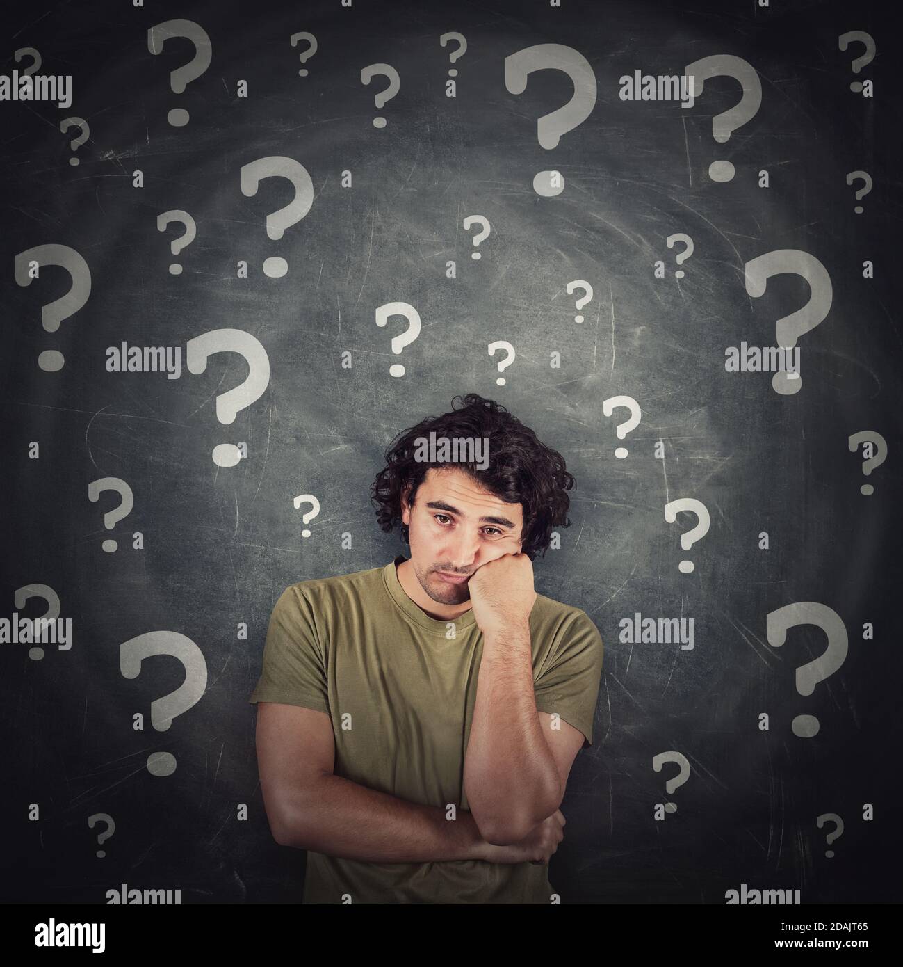 Melancholic young man, long curly hair style, keeps one fist under cheek, looks doubtful and unsure as multiple interrogation marks surrounds him. Puz Stock Photo