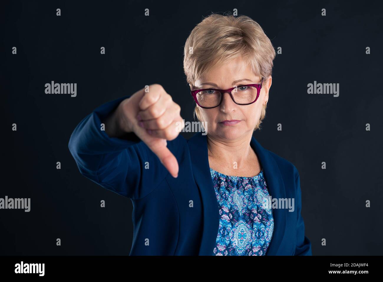 Upset older secretary showing dislike sign on dark background wearing magenta glasees Stock Photo