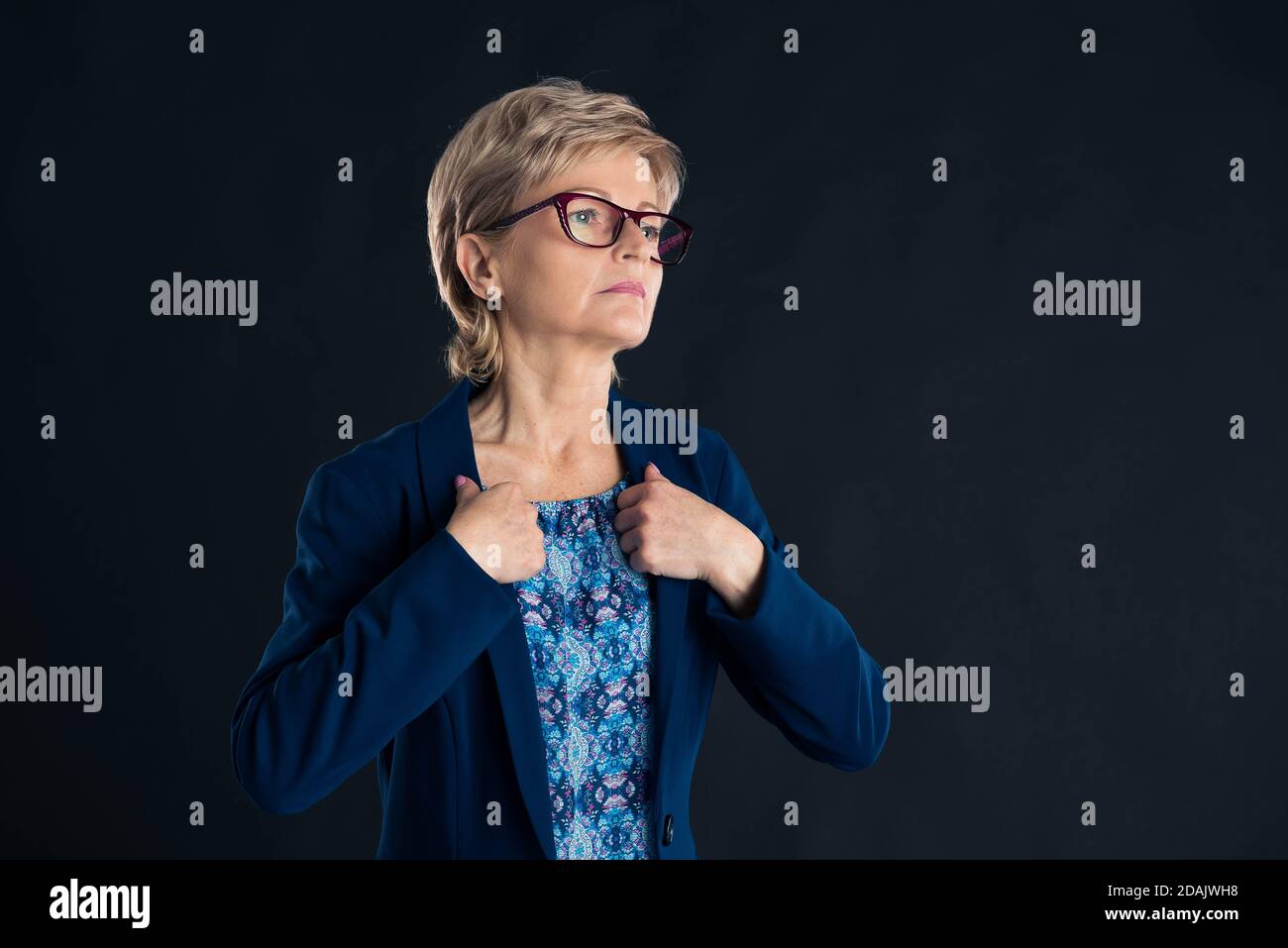 Older secretary fixing her blue jacket on a dark background wearing magenta glasses Stock Photo