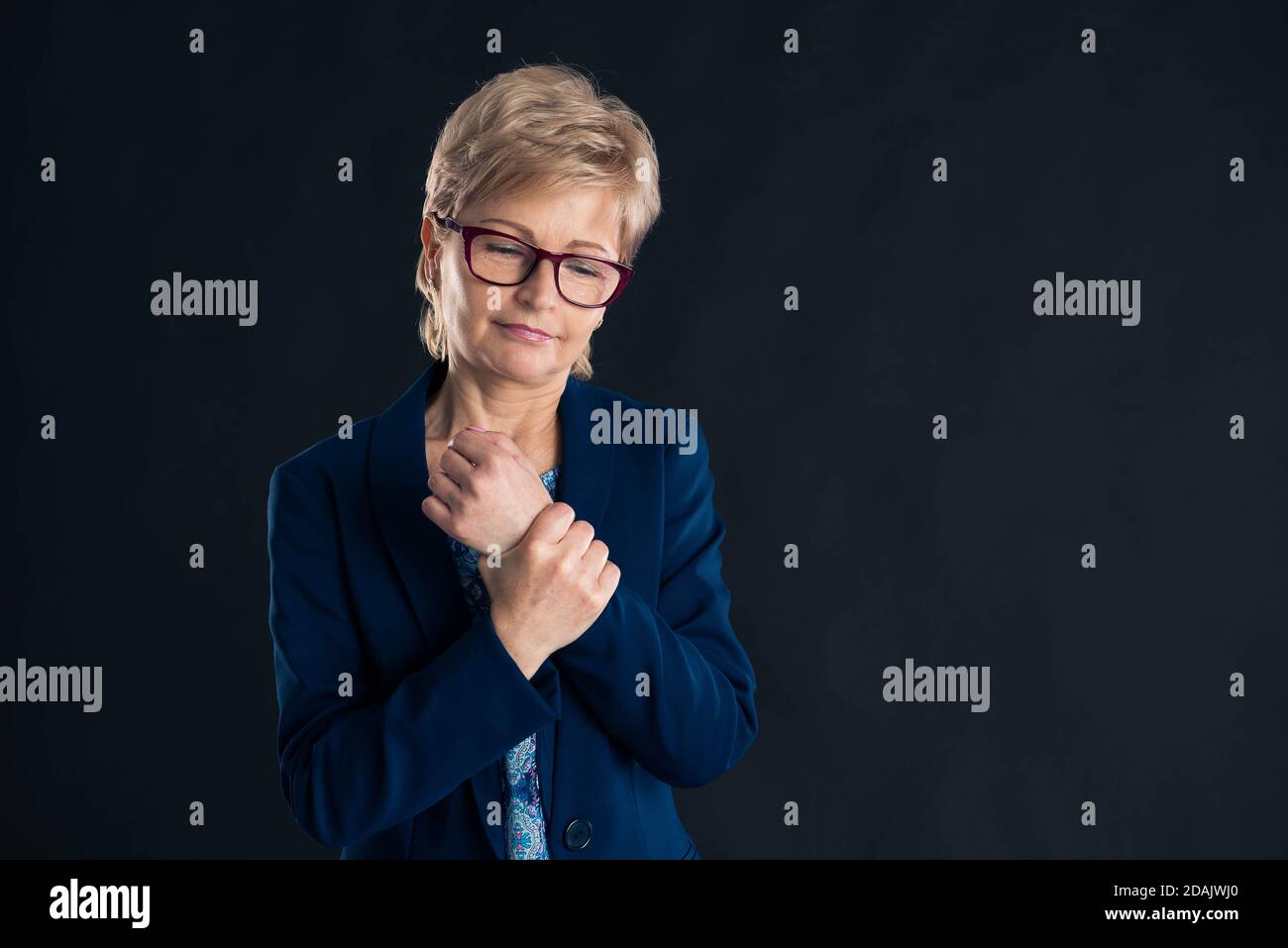 Portrait of an older businesswoman having wrist joint pain wearing a blue jaket on dark background Stock Photo