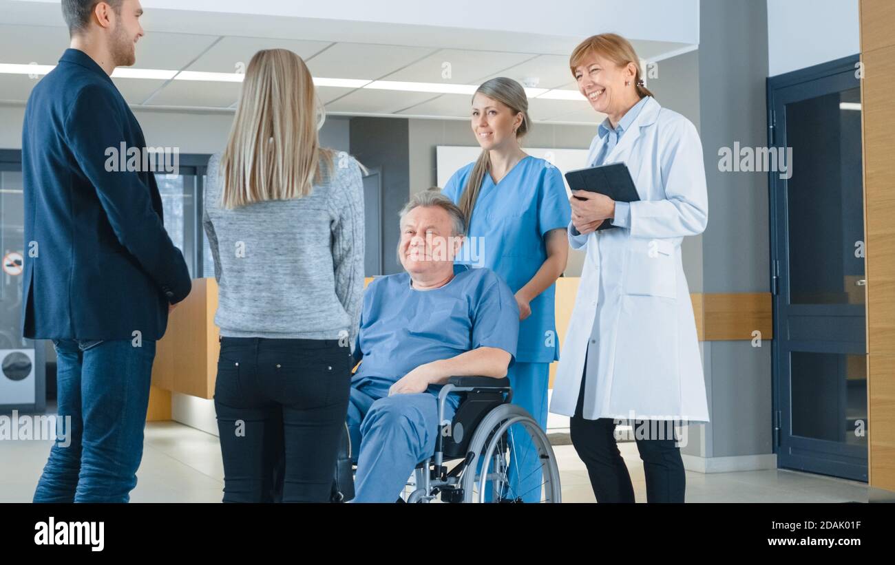 In the Hospital Lobby. Young Couple Visiting Elderly Parent in a Wheelchair, He's aided by Friendly Nurse and Doctor. Happy Family Reunion. New Modern Stock Photo