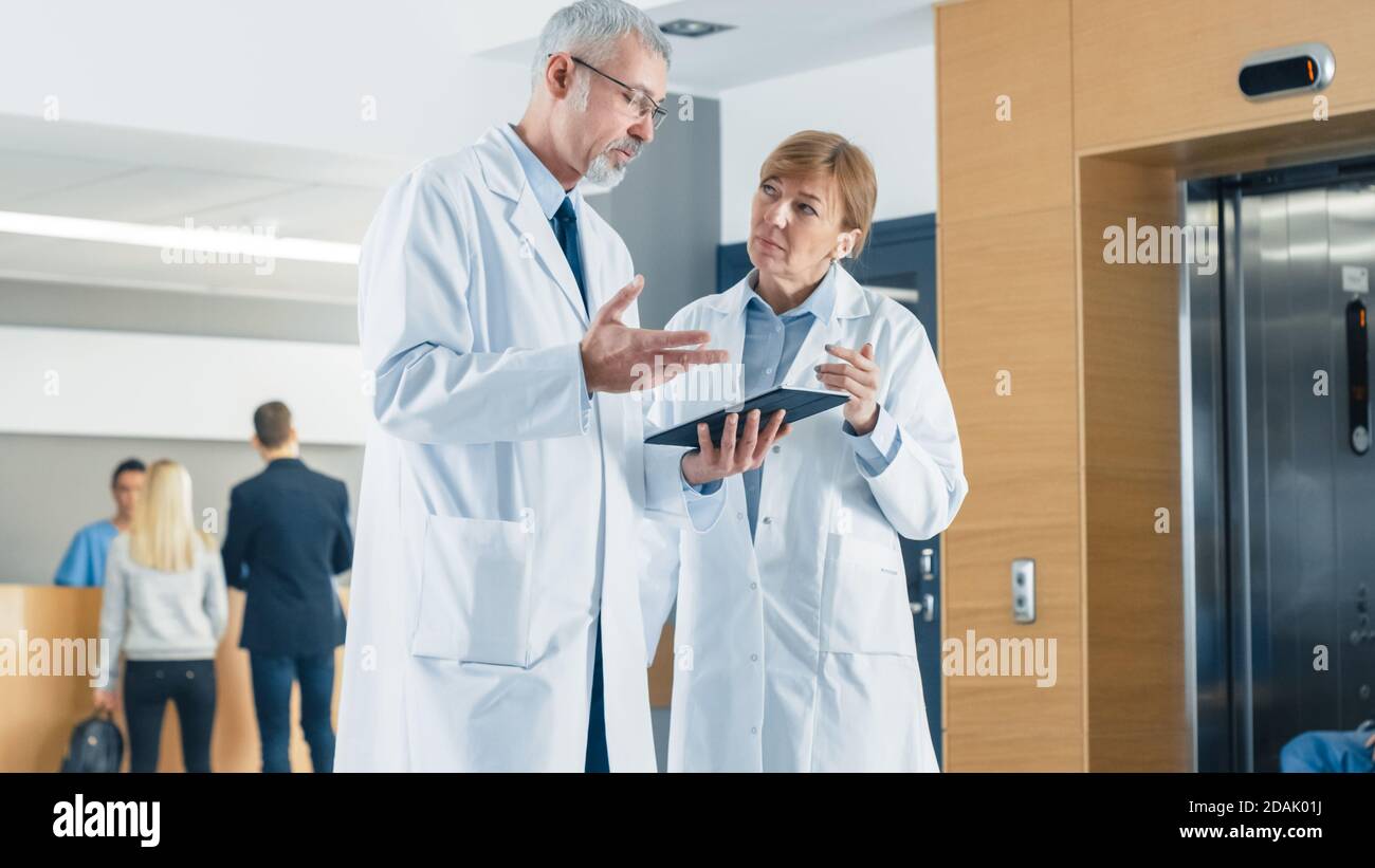 In the Hospital Doctors have Discussion while Using Tablet Computer. In the Background Patients and Medical Personnel. New Modern Fully Functional Stock Photo