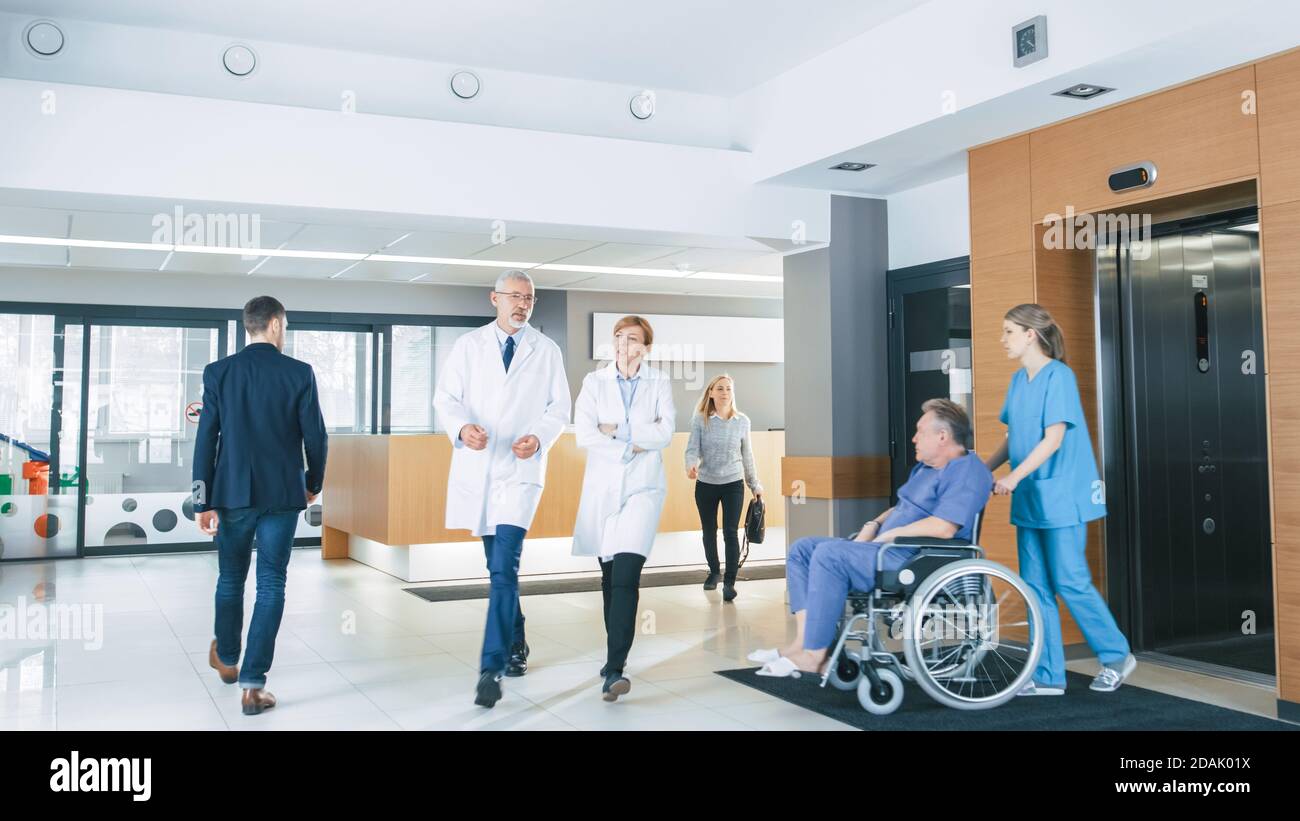 First Floor of the Busy Hospital, Doctors, Nurses and Personnel Busy Working, Assistant Moves Elderly Man in the Wheelchair. New Modern Medical Stock Photo