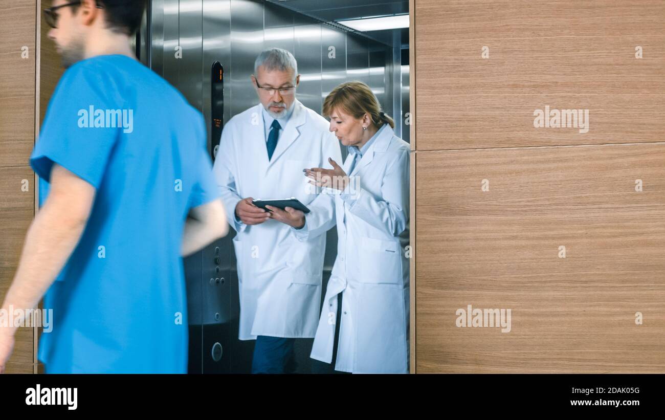 In the Hospital Two Doctors Walk out of the Elevator while have Discussion while Using Tablet Computer. In the Background Patients and Medical Stock Photo