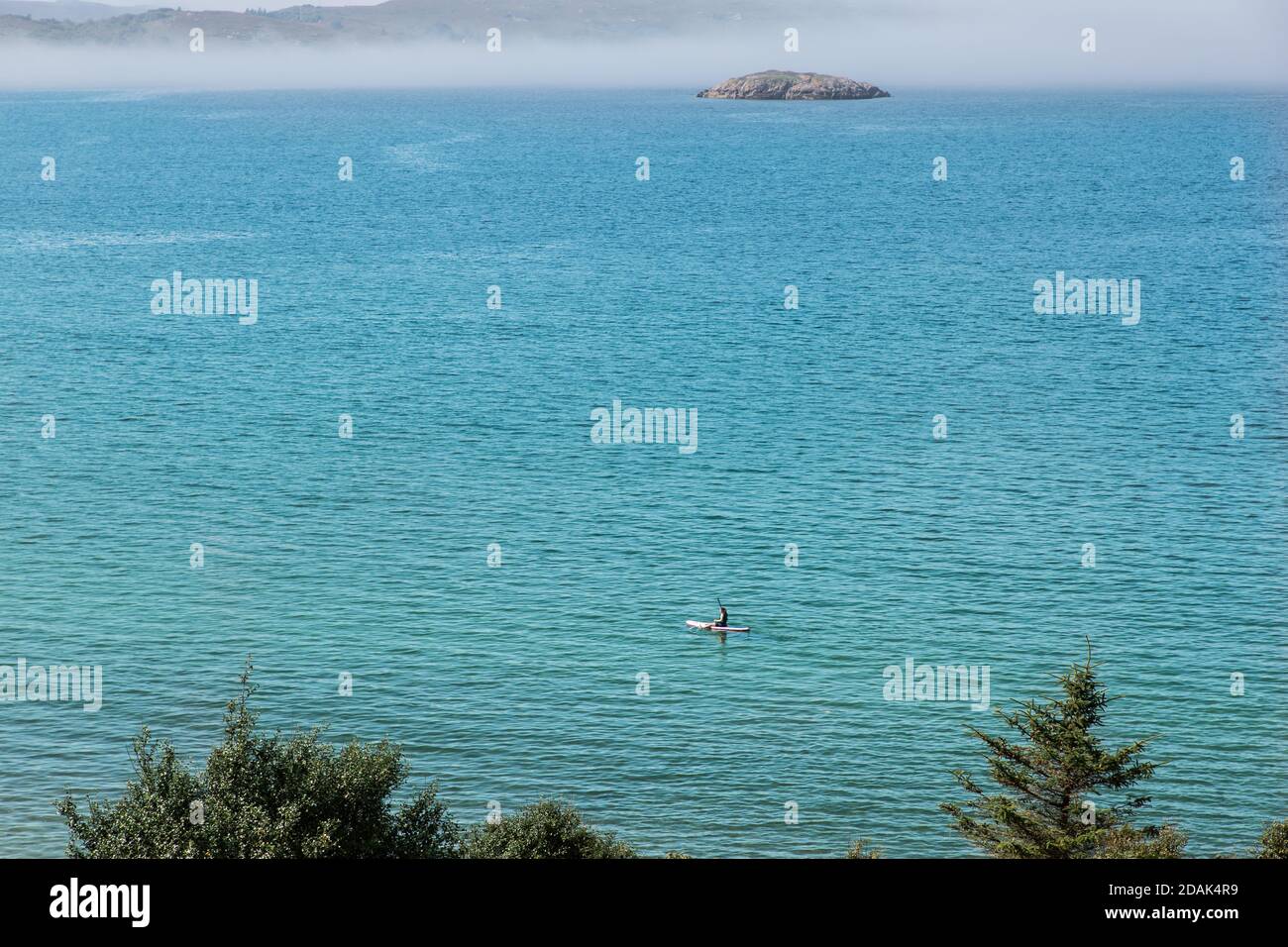 A lone wakeboarder paddles in the turquoise waters of Loch Gairloch in Wester Ross, Scotland. Longa Island is about to be smothered in a sea haar Stock Photo