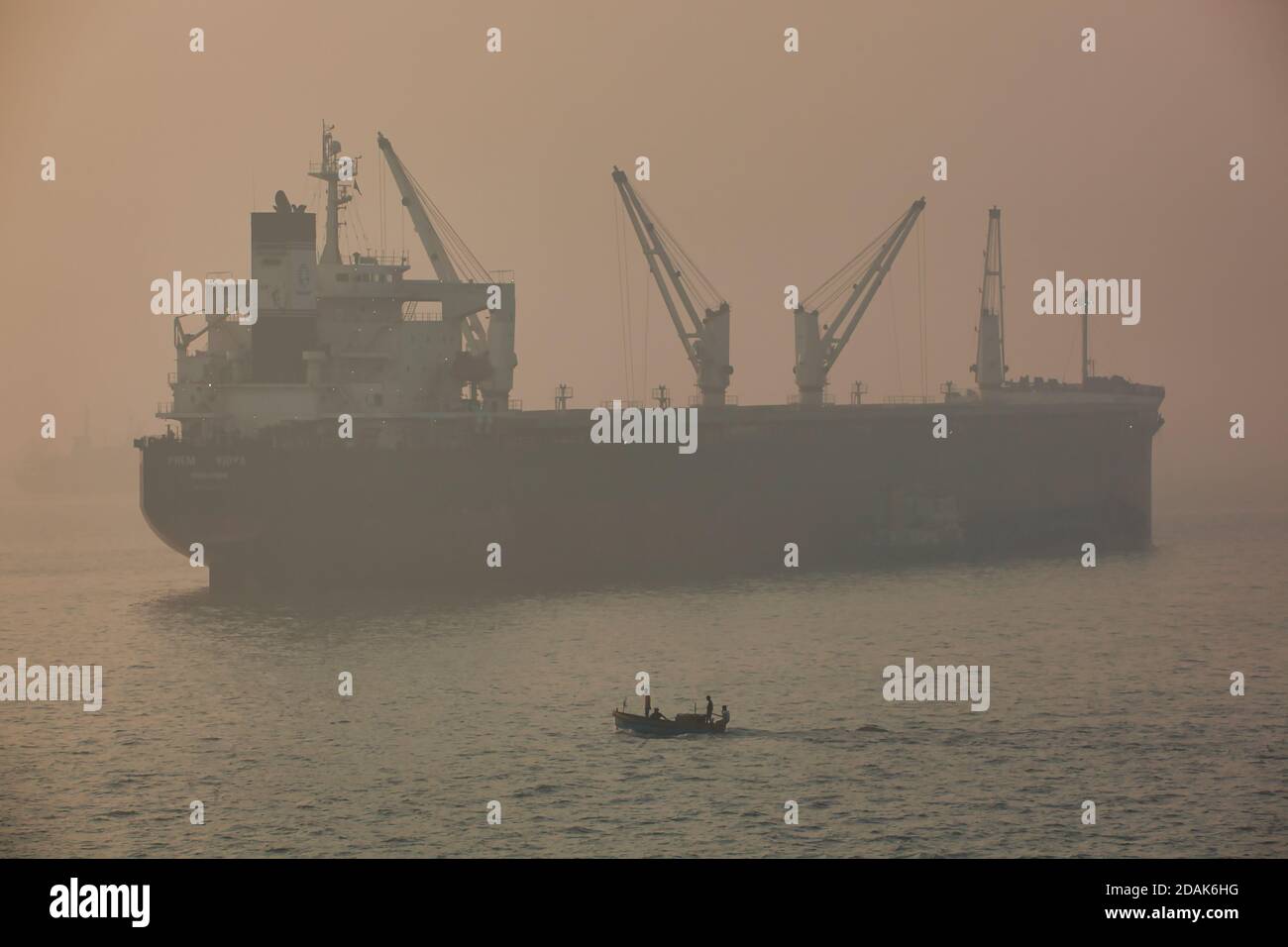 A small motor boat makes it's way across the sea off Mumbai harbour with a cargo ship in the background seen through an early morning haze, India Stock Photo