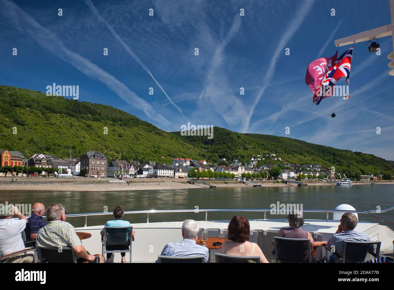 Passengers on a cruise boat on the River Rhine look at the view ahead with many jet vapour trails in the blue sky above. The River Rhine, Germany Stock Photo