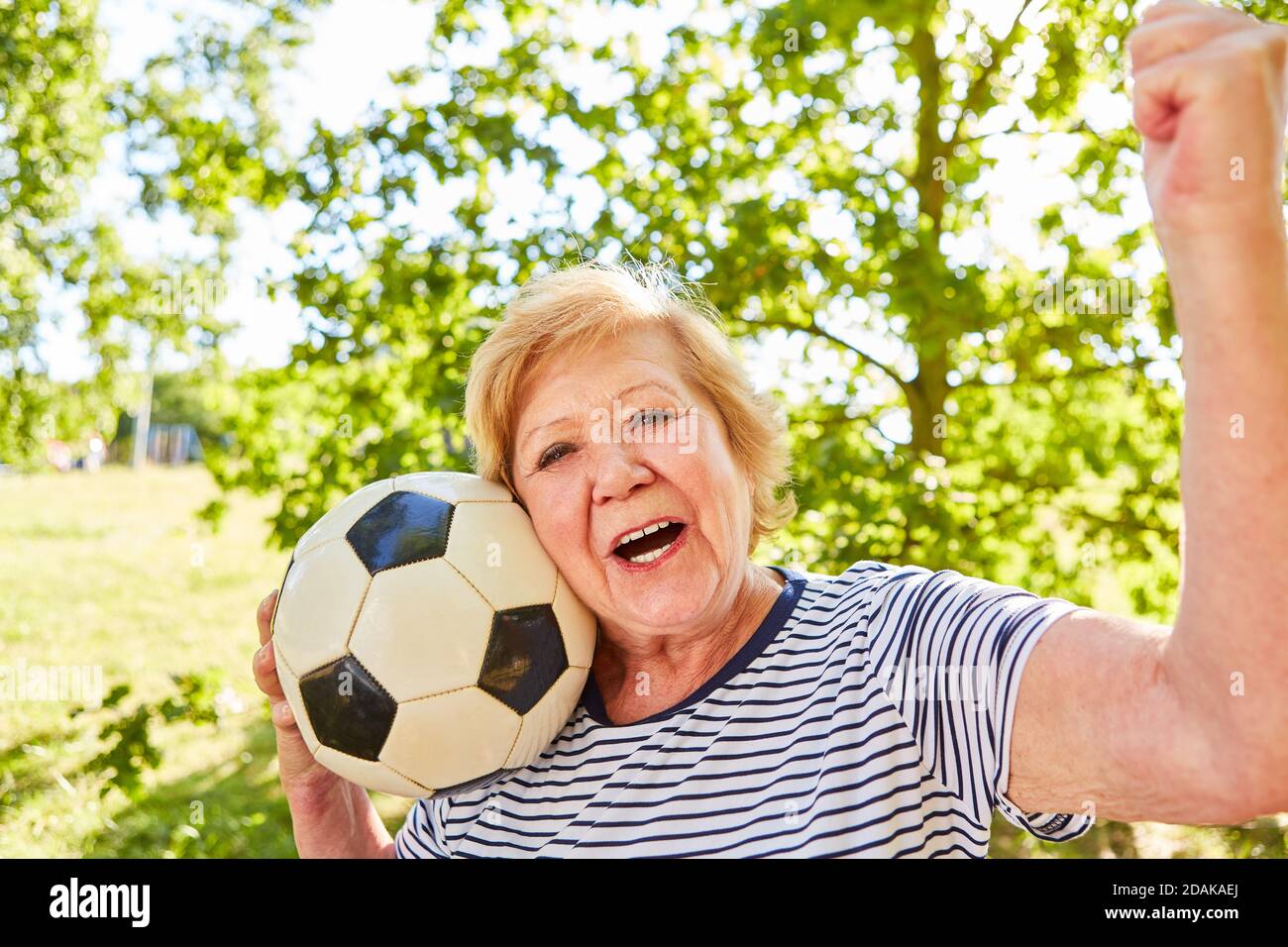Vital elderly woman with soccer ball cheers with clenched fists after the game in the park in summer Stock Photo