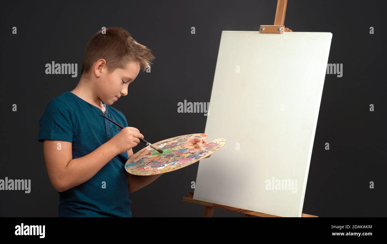 Creative young painter teen boy mixes oil paints on palette while standing in front of blank canvas on easel. Drawing concept. Cut out on gray Stock Photo