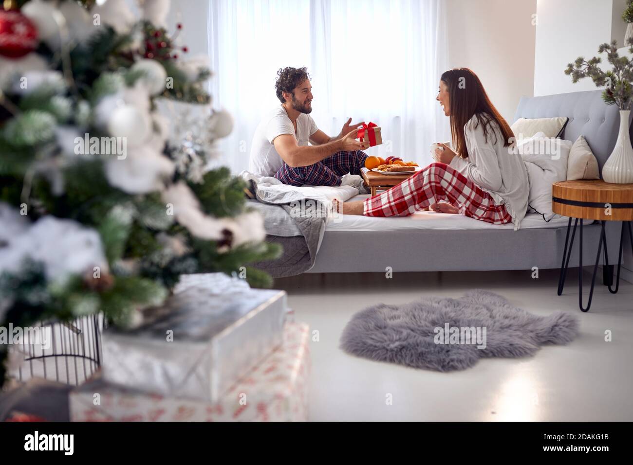 young caucasian beardy guy surprising his girlfriend with Christmas present while having breakfast in bed together Stock Photo
