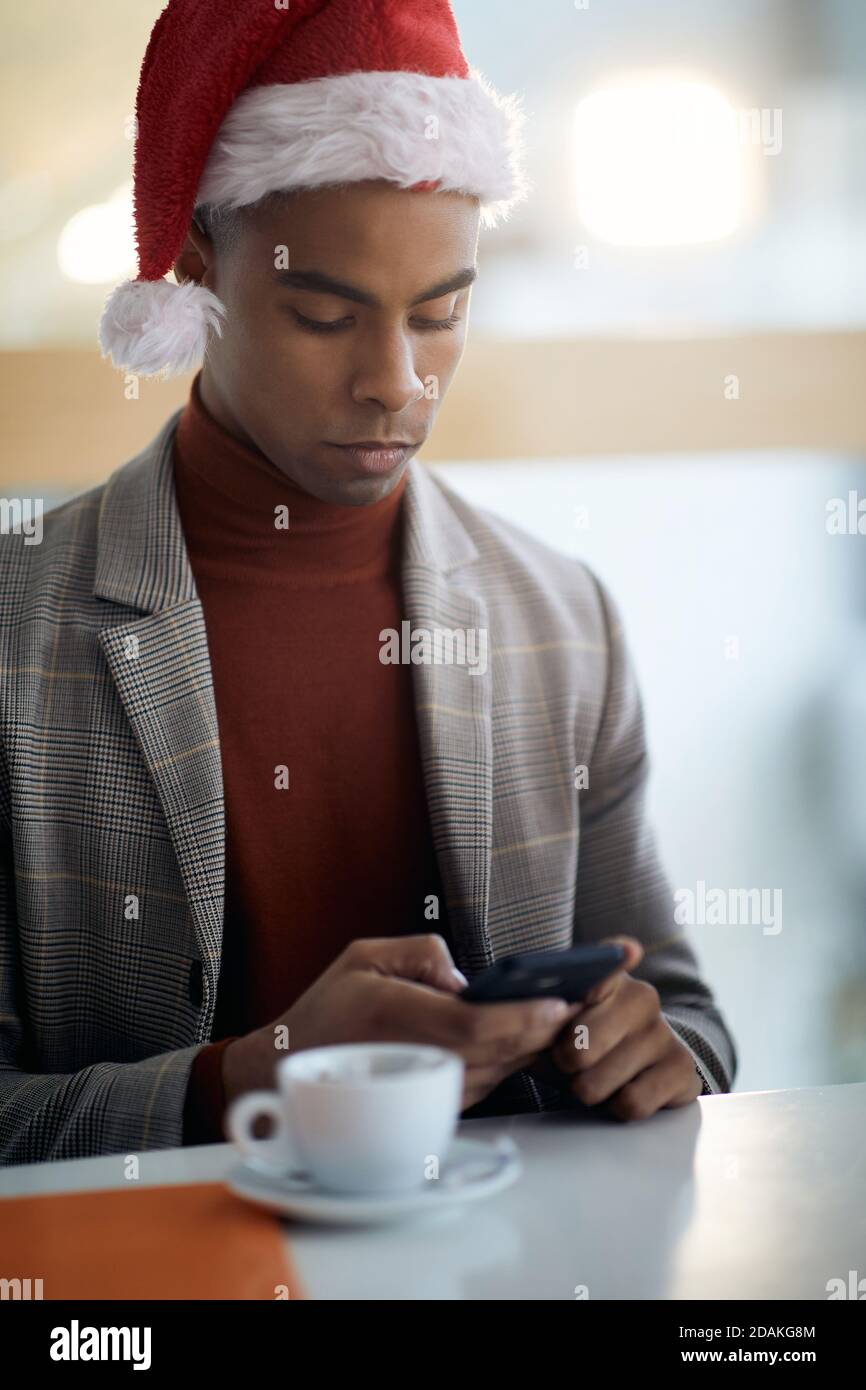 portrait of young afro-american in the office, texting on cell phone, wearing santa hat with cup of coffee in front of him Stock Photo
