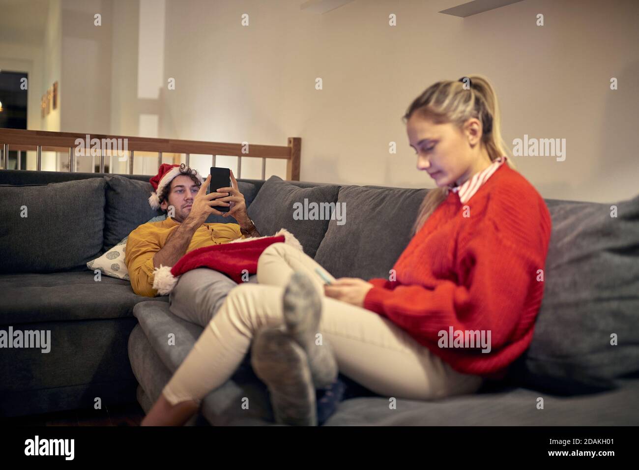 young caucasian male wearing  santa hat using cell phone on christmas laying on sofa with his girlfriend sitting, also using mobile phone Stock Photo