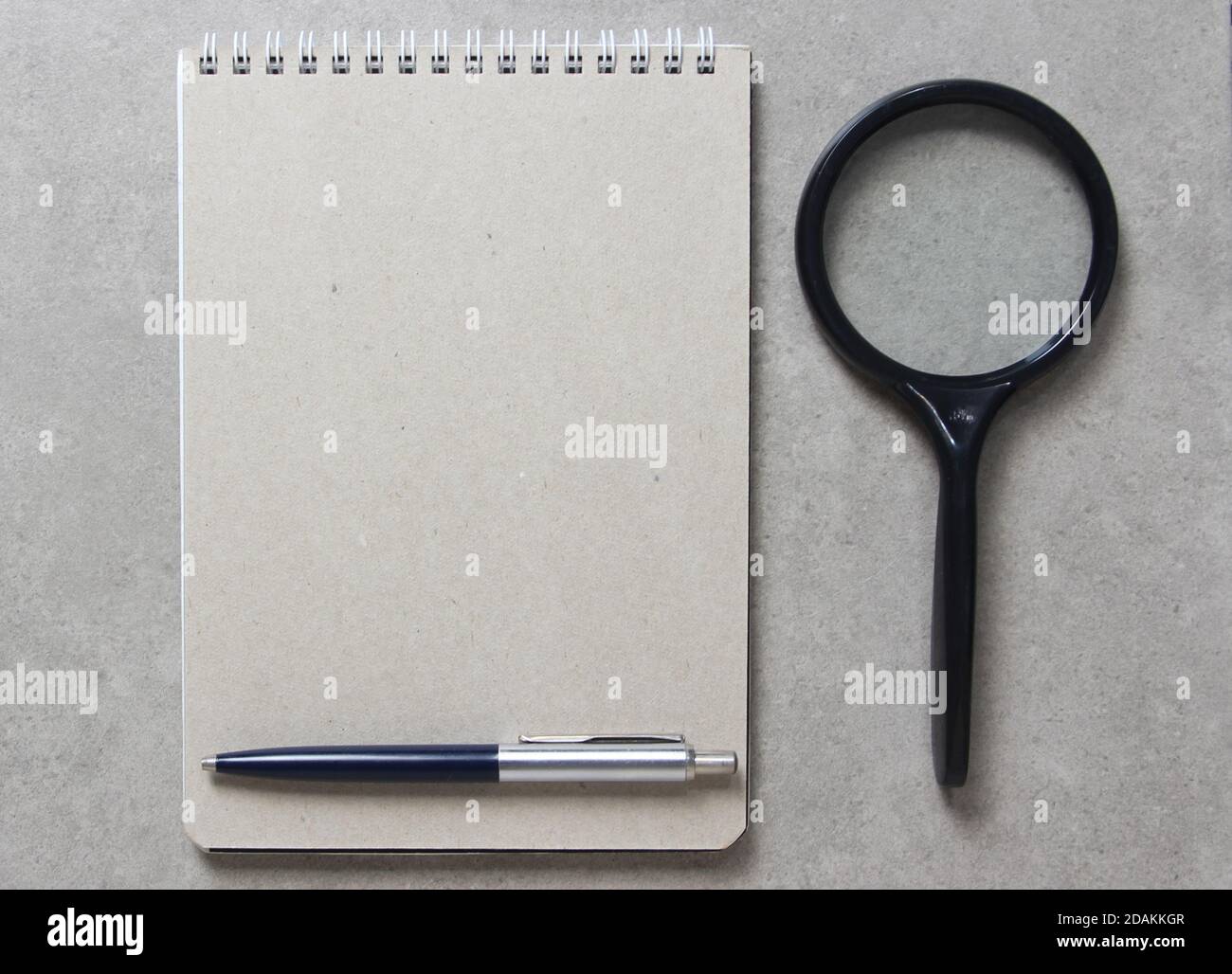 A spring notebook with a sheet of craft paper A5 with a ballpoint pen and black magnifying glass on light grey concrete background. Concept of new idea, business plan and strategy, development and implementation of content. Empty space for text. Stock Photo