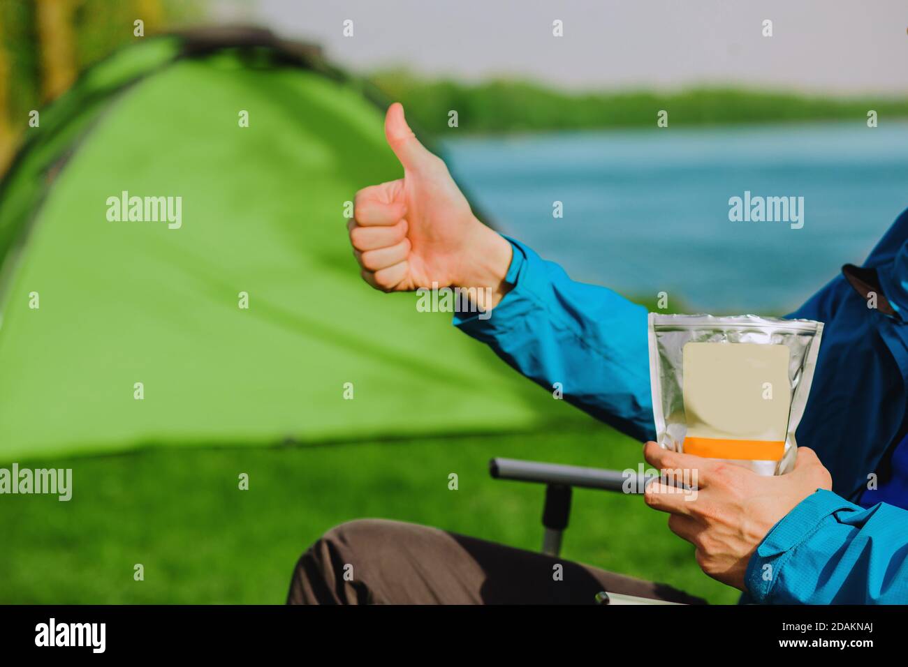 Hand showing thumbs up,enjoy a delicious freeze dried food during the camping holidays. In the background tourist tent and equipment for outdoor activ Stock Photo