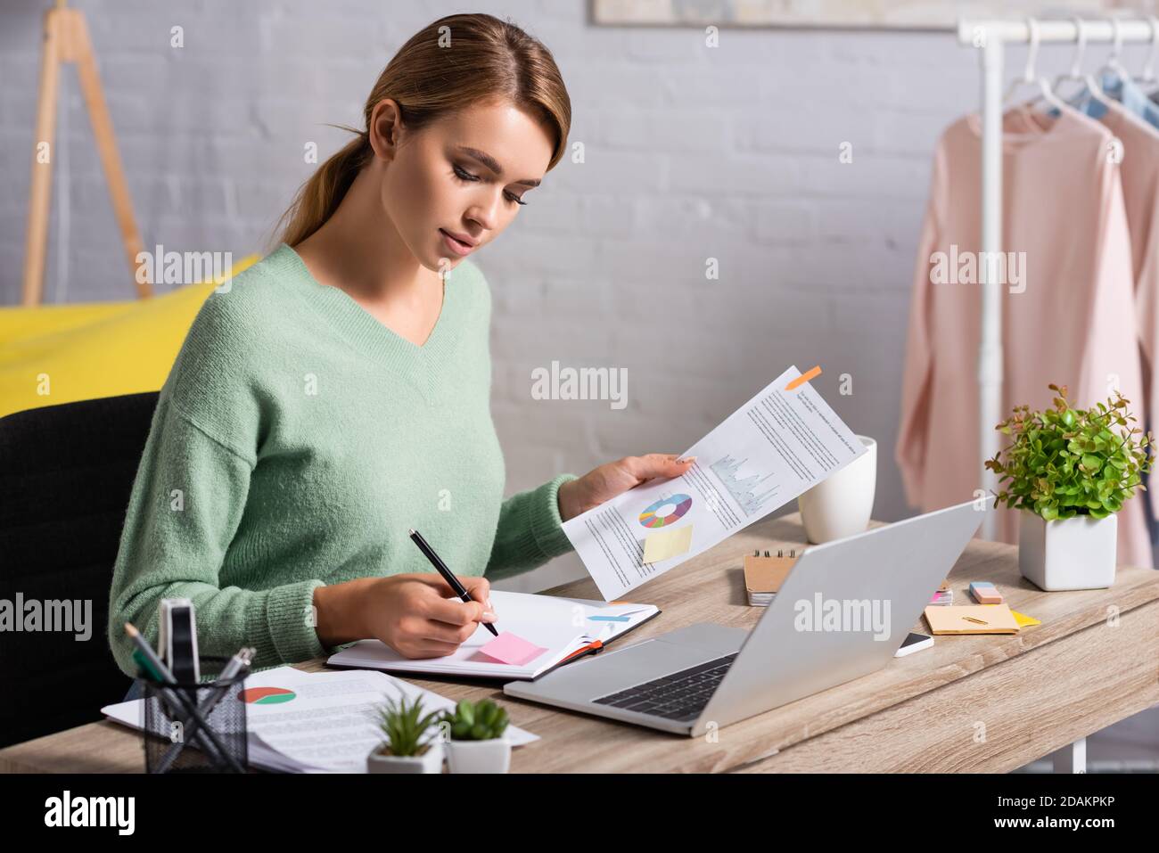 Freelancer writing on notebook while holding document with charts near laptop on table Stock Photo