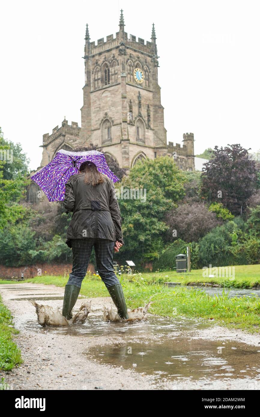 Rear view of isolated woman holding a brolly & splashing into puddles wearing green wellies on a rainy day in summer. Great Britain summer weather. Stock Photo