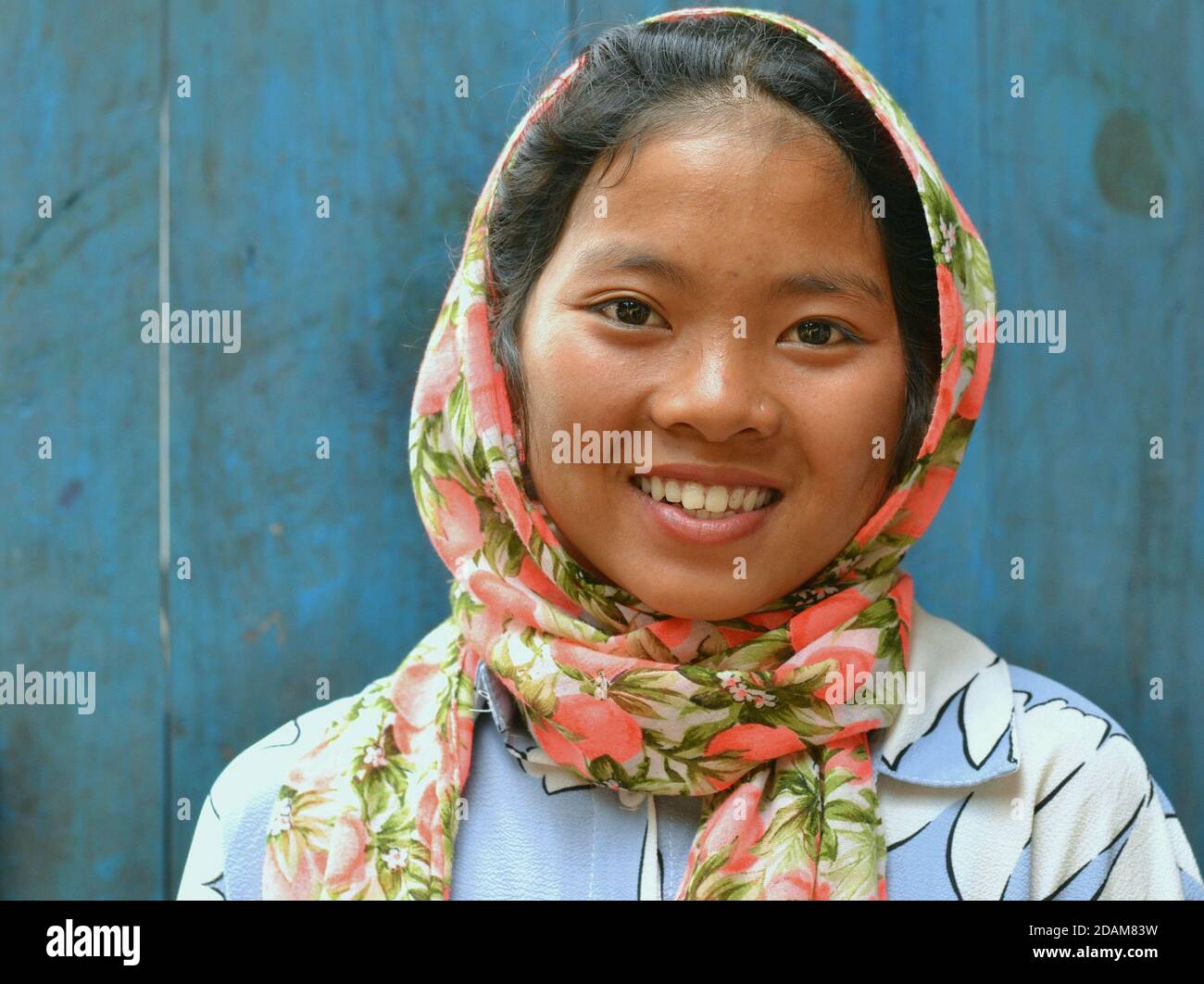 Pretty Northeast Indian Apatani pre-teen girl wears a colorful head scarf and smiles for the camera in front of blue wooden door background. Stock Photo