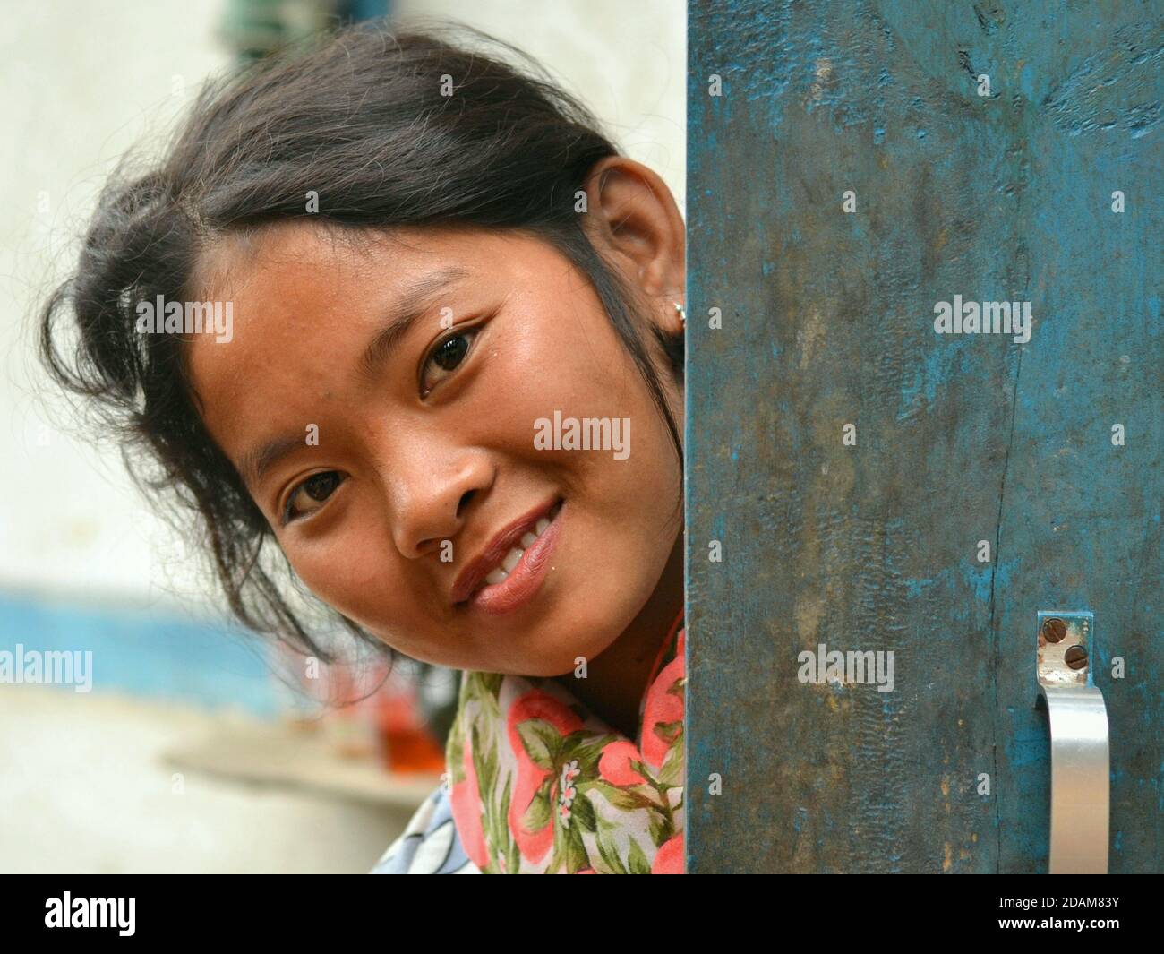 Pretty Northeast Indian Apatani pre-teen girl smiles for the camera and peeks around a blue wooden door. Stock Photo