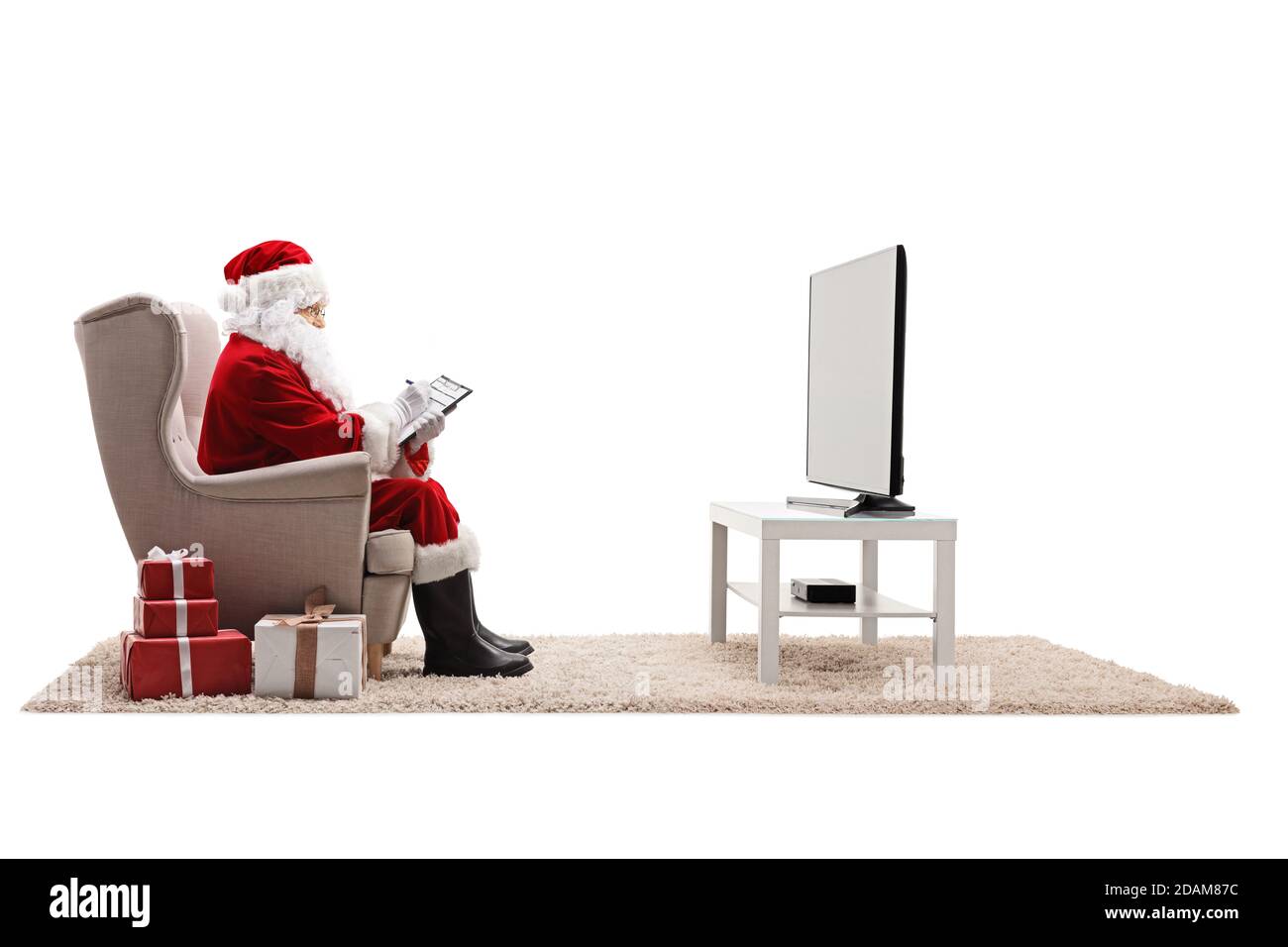 Santa claus sitting in an armchair and writing on a clipboard with presents on the floor isolated on white background Stock Photo