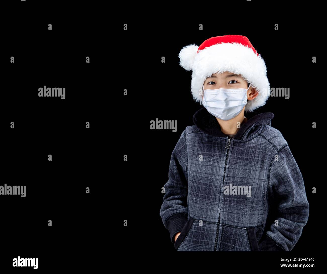 Cute boy during Covid Christmas wearing Santa hat and face mask due to pandamic, isolated on black background with copy space. Stock Photo