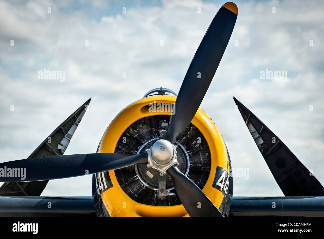 A navy blue World War II aircraft carrier aircraft, with a yellow cowling,  with wings folded against a blue, cloudy sky Stock Photo