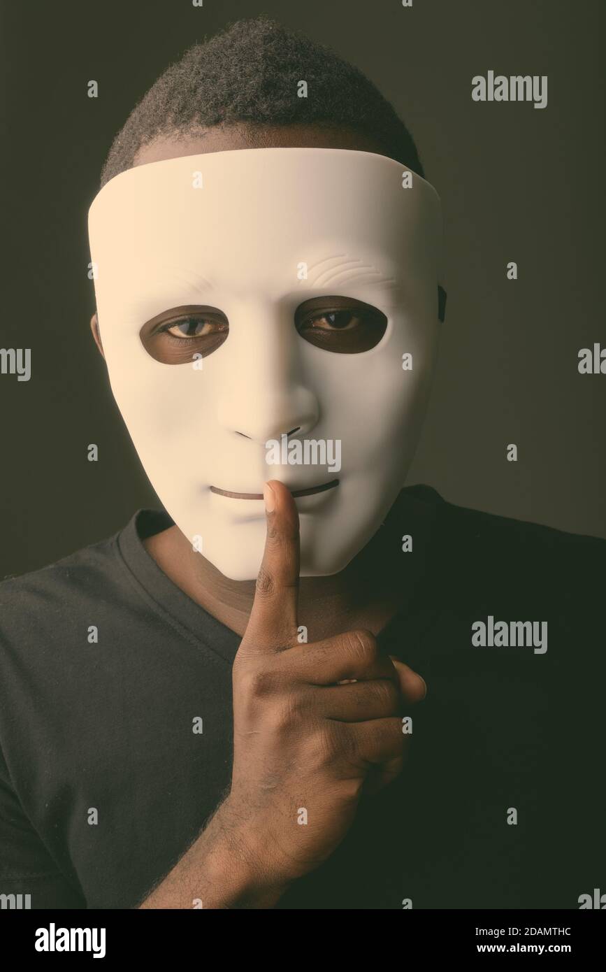 Studio shot of young black African man in dark room wearing mask and holding finger on lips Stock Photo