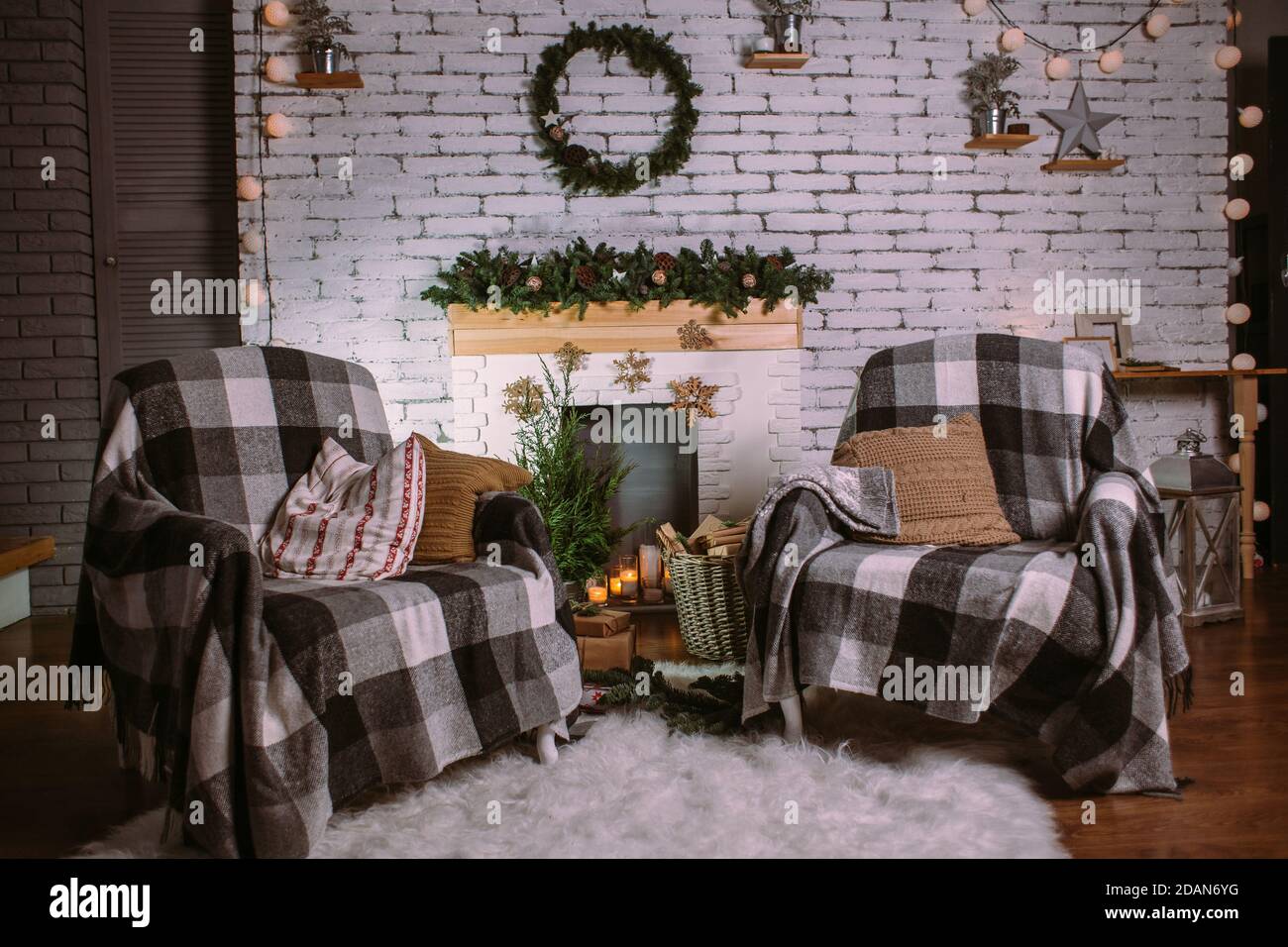 Two armchairs covered with cozy blankets standing near fireplace decorated with garland, candles and basket with gifts. Round green wreath hanging on Stock Photo