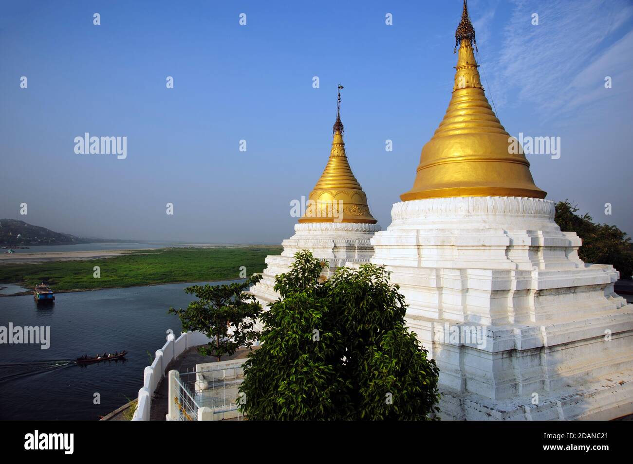 A Burmese boat passes Two gold topped Burmese temple stupas on the banks of the Ayeyarwady river looking across to Sagaing near Mandalay Myanmar Stock Photo