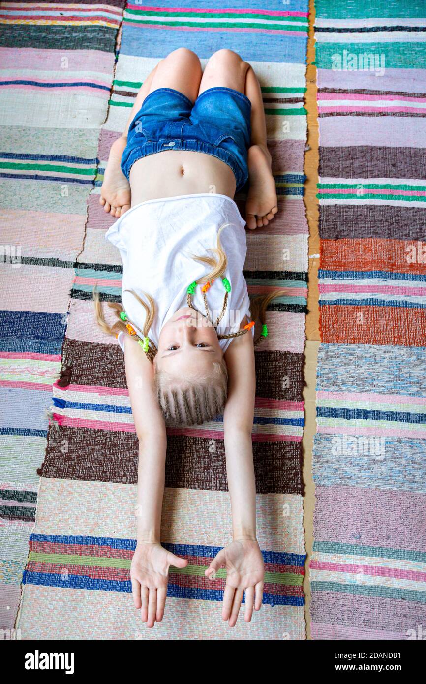 Caucasian girl with braids in a white T-shirt is doing yoga, stretching at home on a striped fabric rug.Digital detox, reunion with nature, healthy br Stock Photo