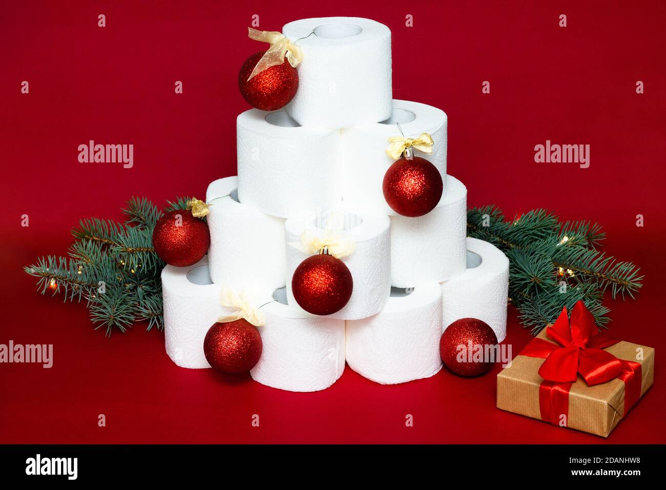 Christmas tree made from rolls of toilet paper, decorated with shiny balls, near a gift and fir branches on a red background Stock Photo