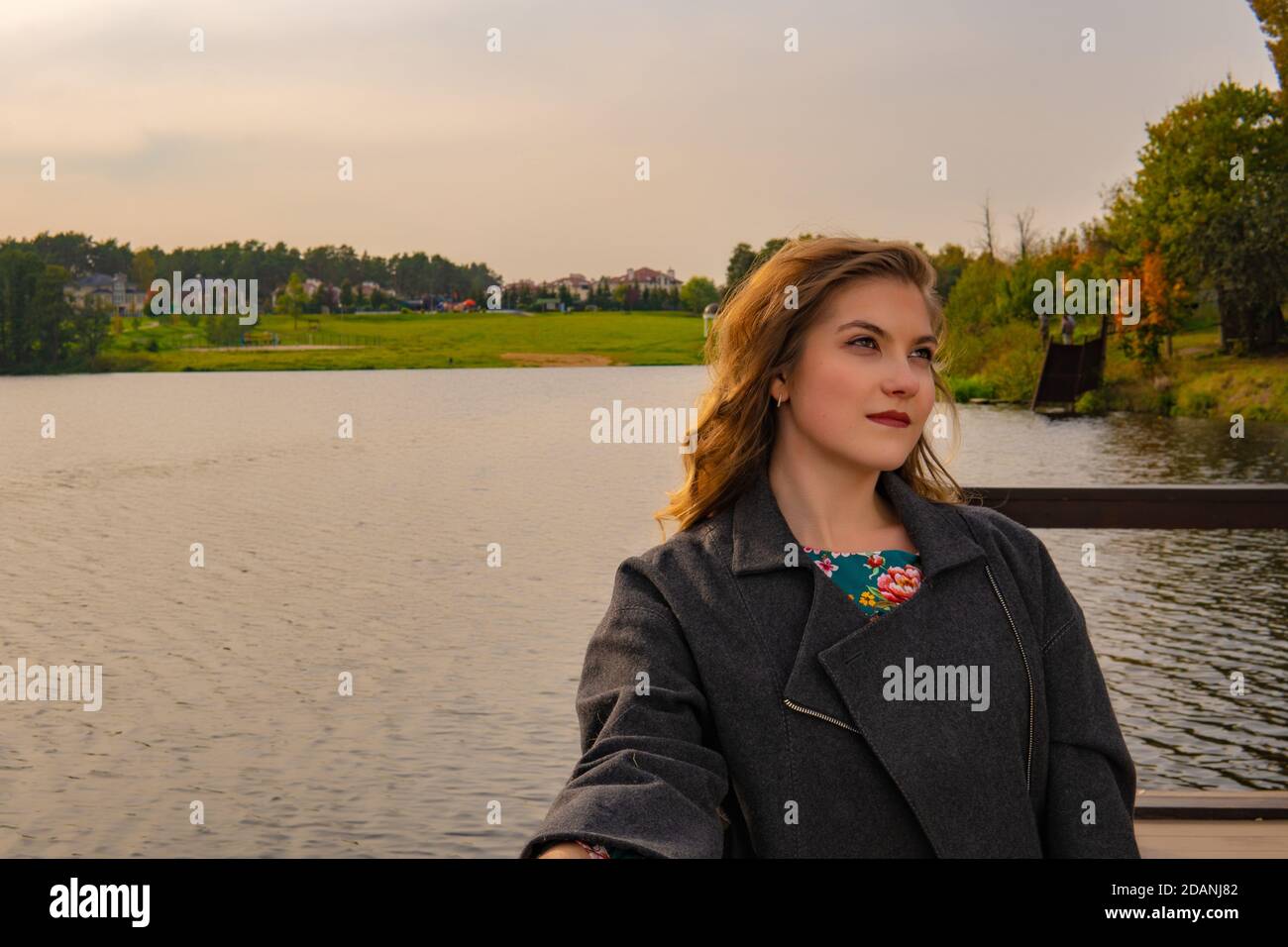 Cheerful adult woman standing near the lake with a charismatic appearance with a Caucasian appearance in a coat in autumn on the pier in the afternoon Stock Photo